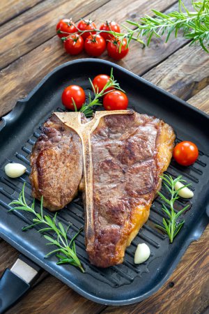 Photo for T-bone steak on frying grill pan with cherry tomatoes, garlic and rosemary on wooden table - Royalty Free Image