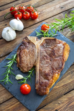 Photo for T-bone steak on frying grill pan with cherry tomatoes, garlic and rosemary on wooden table - Royalty Free Image