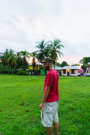 Foto de Hombre con barba y sombrero de pie frente a viviendas tropicales. - Imagen libre de derechos