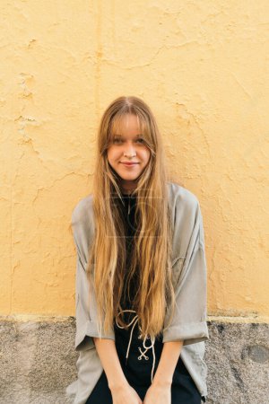 Foto de Joven rubia adulta con el pelo largo sola al aire libre de pie contra una pared amarilla mirando a la cámara. - Imagen libre de derechos