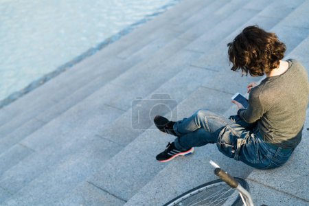 Photo for Woman resting from a bike ride texting on her phone sitting on stairs outdoors. - Royalty Free Image
