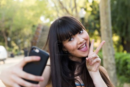 Foto de Bonita mujer europea asiática toma una selfie sonriendo al smartphone en las calles de Barcelona con luz natural de un día soleado - Imagen libre de derechos