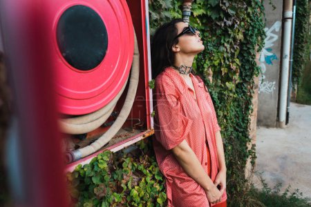 Photo for Young woman with cool black sunglasses wearing red latex pants and pink shirt posing to camera with a wall with vegetation and fire hose in the background. - Royalty Free Image