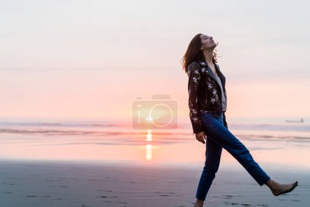 Foto de Adulto joven caminando por la playa con los ojos cerrados mirando hacia arriba disfrutando de una puesta de sol dorada - Imagen libre de derechos