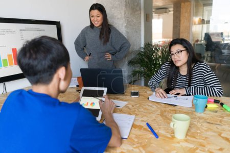 Foto de Mesa de oficina con tres personas asiáticas sentadas y usando dispositivos mientras trabajan con gráficos. - Imagen libre de derechos