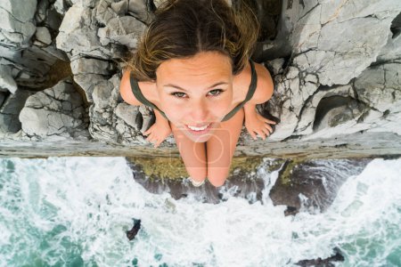 Foto de Fotografía tomada desde arriba de una joven rubia sentada en el borde de un acantilado mirando hacia la cámara. - Imagen libre de derechos