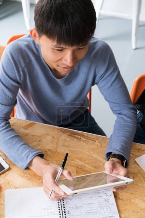 Foto de Asiático macho sentado en la mesa con tableta y bloc de notas de trabajo. - Imagen libre de derechos