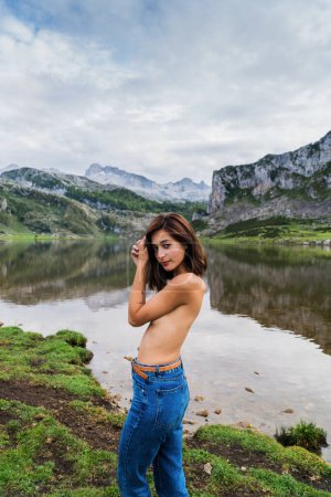 Foto de Foto lateral de una joven morena en topless usando jeans al aire libre en la naturaleza mirando a la cámara que cubre sus pechos con un paisaje de lago y montañas en el fondo. - Imagen libre de derechos