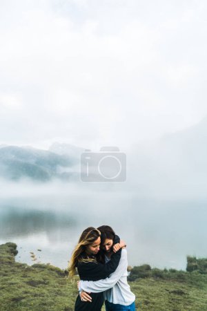 Foto de Pareja femenina de jóvenes adultos abrazándose en un paisaje natural increíble con niebla y nubes en el fondo. - Imagen libre de derechos