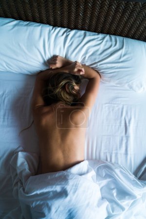 Téléchargez les photos : Tourné d'en haut d'une femme méconnaissable couchée face contre terre nue recouverte de draps de lit. - en image libre de droit