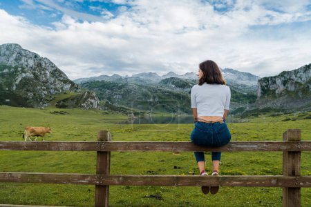 Foto de Mujer joven sentada en una valla disfrutando de una vista al lago con altas montañas. - Imagen libre de derechos