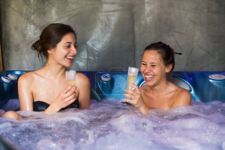 Foto de Dos parejas femeninas en una bañera de hidromasaje burbujeante haciendo un brindis con champán. - Imagen libre de derechos