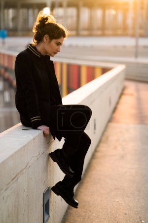 Téléchargez les photos : Jeune adulte assis sur une rampe de pont regardant vers le bas portant tout noir - en image libre de droit