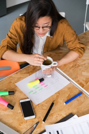 Foto de Joven mujer china sentada en el escritorio con taza de té y mirando el papel. - Imagen libre de derechos