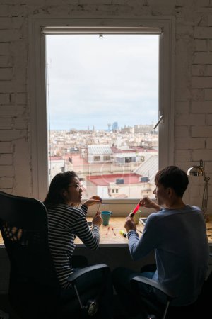 Téléchargez les photos : Vue arrière de deux collègues assis sur le bureau près de la fenêtre prenant un café et parlant. - en image libre de droit