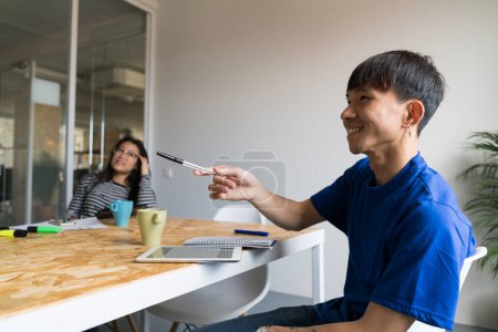 Téléchargez les photos : Vue latérale du jeune homme asiatique assis à table avec tablette et bloc-notes pointant du doigt et souriant. - en image libre de droit