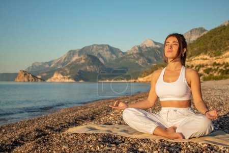 Photo for A girl with curly hair in a white tracksuit, sitting in the lotus position with closed eyes and meditating, against the background of mountains, sea and sand - Royalty Free Image