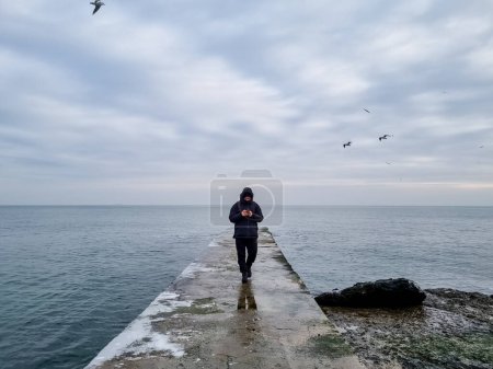 Foto de Un hombre camina solo en el muelle y mira el teléfono, en ropa de invierno, el mar es áspero, el tiempo lluvioso, las gaviotas están volando - Imagen libre de derechos