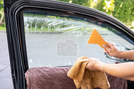 Photo for Auto specialist worker hand using squeegee blade or spatula and cloth wipe cleaning old car window film tint after spraying soapy solution. Car side window film removal and tinting installation - Royalty Free Image
