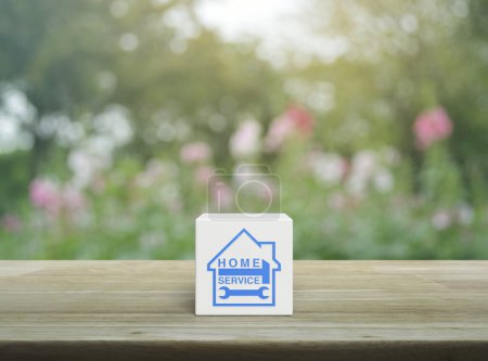 Martillo y llave inglesa con icono de la casa en el cubo de bloque blanco en la mesa de madera sobre la flor rosa borrosa y el árbol en el jardín, concepto de servicio a domicilio de negocios