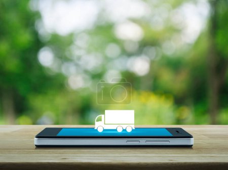 Icône plate de camion sur l'écran moderne de téléphone portable intelligent sur la table en bois sur l'arbre vert flou dans le parc, concept de service en ligne de transport d'affaires