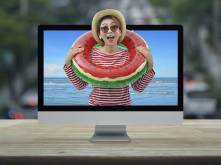 Happy young asian woman wearing red stripped shirt and straw hat in watermelon inflatable ring with tropical sea on computer screen on table over blur of rush hour with cars and road, Business summer holiday online concept