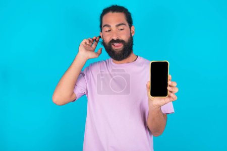 Photo for Photo of nice pretty Caucasian man with beard wearing violet T-shirt over blue background demonstrate phone screen hold hair tails - Royalty Free Image