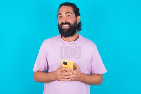 Foto de Foto de hombre caucásico loco asombrado con barba usando camiseta violeta sobre fondo azul mantenga el concepto de retroalimentación de disgusto del teléfono inteligente - Imagen libre de derechos