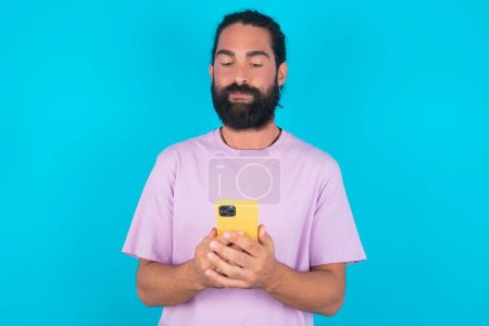 Téléchargez les photos : Portrait d'homme caucasien confiant sérieux avec barbe portant T-shirt violet sur fond bleu tenant le téléphone dans les deux mains - en image libre de droit