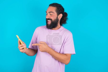 Foto de Retrato lindo hombre caucásico asombrado con barba vistiendo camiseta violeta sobre fondo azul impresionado increíble consejo increíble notificación increíble - Imagen libre de derechos