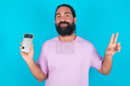 Téléchargez les photos : Homme caucasien avec barbe portant T-shirt violet sur fond bleu tenant dispositif moderne montrant v-signe - en image libre de droit