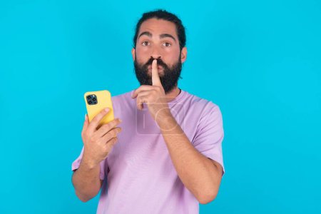 Foto de Hombre caucásico con barba vistiendo camiseta violeta sobre fondo azul sosteniendo gadget moderno pedir no contar secretos - Imagen libre de derechos