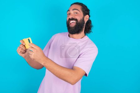 Téléchargez les photos : Joli accro gai homme caucasien avec barbe portant T-shirt violet sur fond bleu en utilisant gadget jouer jeu de réseau - en image libre de droit