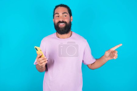 Foto de Hombre caucásico asombrado con barba vistiendo camiseta violeta sobre fondo azul sosteniendo su teléfono y señalando con el dedo a un lado en el espacio de copia vacío - Imagen libre de derechos