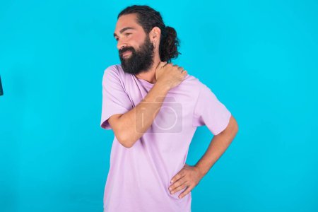 Foto de Caucásico hombre con barba vistiendo violeta camiseta sobre fondo azul tiene dolor de espalda - Imagen libre de derechos
