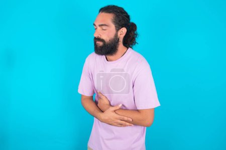 Foto de Caucásico hombre con barba usando violeta camiseta sobre fondo azul tiene dolor de estómago - Imagen libre de derechos