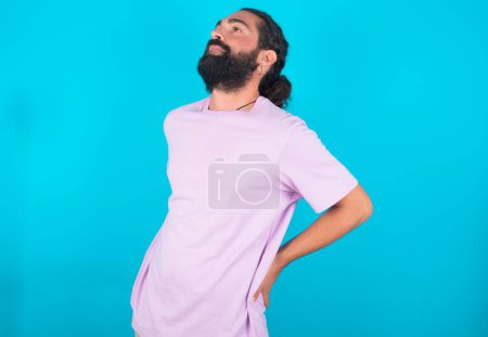 Foto de Caucásico hombre con barba vistiendo violeta camiseta sobre fondo azul tiene dolor de espalda - Imagen libre de derechos