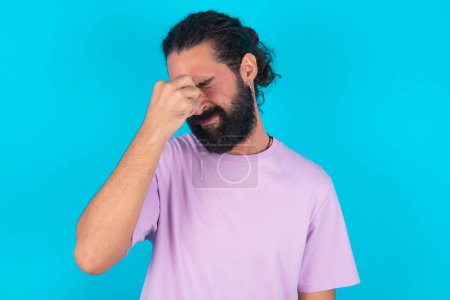 Téléchargez les photos : Homme caucasien triste avec barbe portant T-shirt violet sur fond bleu souffrant de maux de tête tenant la main sur son visage - en image libre de droit