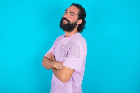 Foto de Retrato de hombre caucásico con barba vistiendo camiseta violeta sobre fondo azul de pie con los brazos cruzados y sonriendo - Imagen libre de derechos