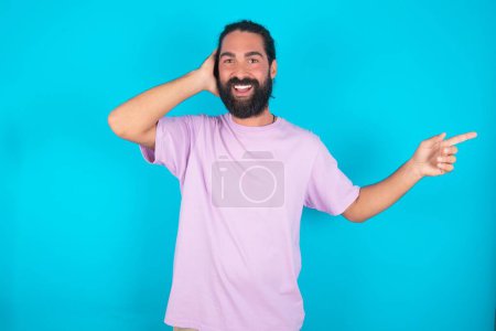 Téléchargez les photos : Surpris homme caucasien avec barbe portant T-shirt violet sur fond bleu pointant vers l'espace vide tenant la main sur la tête - en image libre de droit