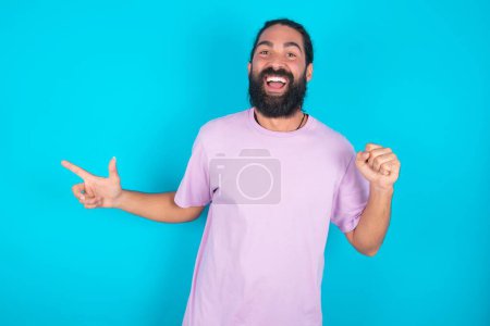 Photo for Caucasian man with beard wearing violet T-shirt over blue background points at empty space holding fist up, winner gesture. - Royalty Free Image