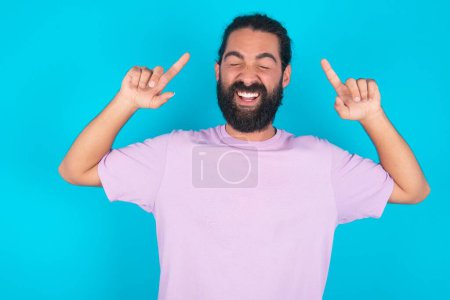Foto de Foto de hombre caucásico loco con barba vistiendo camiseta violeta sobre fondo azul gritando y señalando con los dedos a los ojos cerrados pelo - Imagen libre de derechos