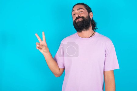 Photo for Caucasian man with beard wearing violet T-shirt over blue background makes peace gesture keeps lips folded shows v sign. Body language concept - Royalty Free Image