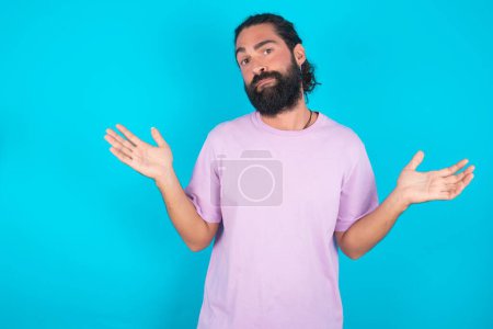 Téléchargez les photos : Insouciant homme caucasien attrayant avec barbe portant T-shirt violet sur fond bleu haussant les épaules, oups. - en image libre de droit
