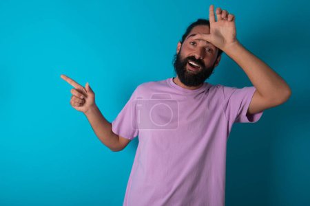 Foto de Hombre caucásico con barba vistiendo camiseta violeta sobre fondo azul mostrando signo perdedor y apuntando al espacio vacío - Imagen libre de derechos