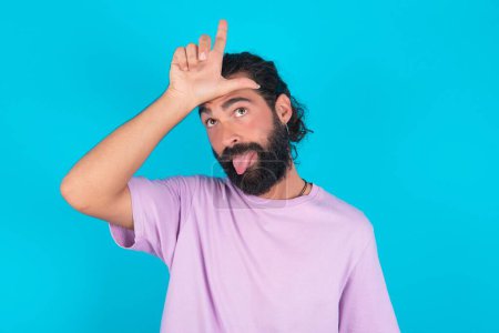 Foto de Divertido hombre caucásico con barba vistiendo camiseta violeta sobre fondo azul hace gesto perdedor burlándose de alguien sobresale lengua haciendo mueca cara. - Imagen libre de derechos