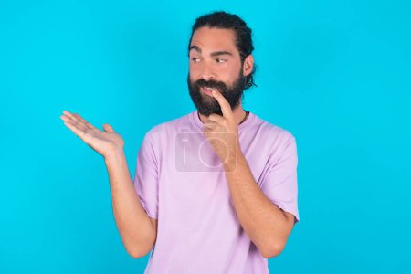 Téléchargez les photos : Positif homme caucasien avec barbe portant T-shirt violet sur fond bleu annonce promo touche dents avec le doigt. - en image libre de droit