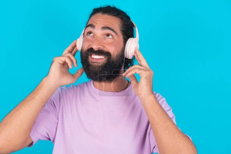 Téléchargez les photos : Homme caucasien joyeux avec barbe portant T-shirt violet sur fond bleu chante chanson garde la main près de la bouche comme si le microphone écoute playlist préférée via casque - en image libre de droit