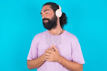 Photo for Caucasian man with beard wearing violet T-shirt over blue background  wears stereo headphones listening to music concentrated and looking aside with interest. - Royalty Free Image