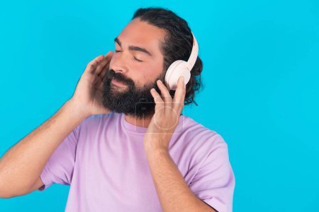 Téléchargez les photos : Homme caucasien barbu portant un T-shirt violet sur fond bleu avec un casque sur la tête, écoute de la musique, apprécie la chanson préférée avec les yeux fermés, tenant la main sur un casque. - en image libre de droit